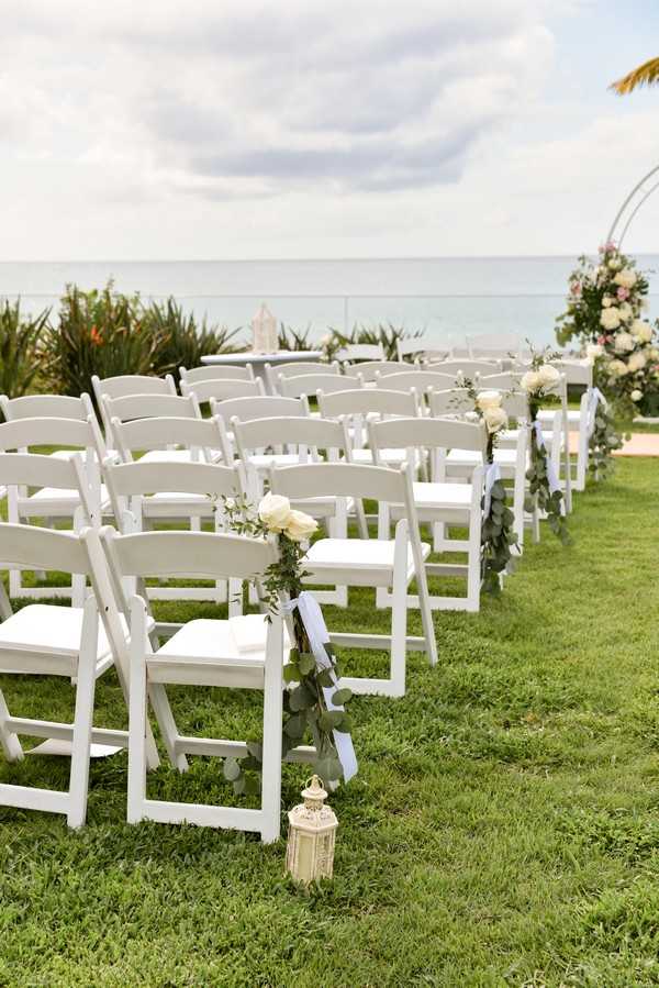 An outdoor wedding ceremony setup on a lawn overlooking the ocean, with no guests or couple present. Rows of white folding chairs with white cushions are arranged on either side of a central aisle, each aisle chair decorated with a small arrangement of cream roses, eucalyptus greenery, and a dusty blue ribbon, with cream lattice lanterns placed on the grass at the base of each aisle chair. In the background, a circular arch decorated with blush, cream, and green floral arrangements marks the altar area. The overall decor palette is white, cream, soft blush, and dusty blue with greenery accents, giving a classic coastal style. Wide shot taken from a low angle along the aisle.