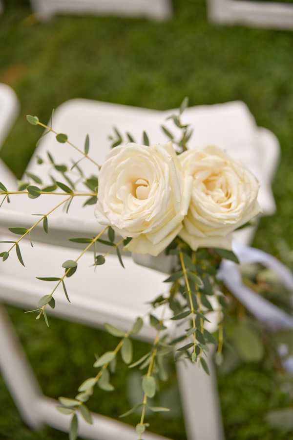 Close-up detail shot of an outdoor ceremony aisle marker featuring two cream roses paired with trailing eucalyptus stems, attached to a white folding chair with a white ribbon. Additional white folding chairs are visible in soft focus in the background, set on grass. The floral styling is simple and classic, with a white and green color palette. The composition is a tight detail shot focused on the chair decoration.