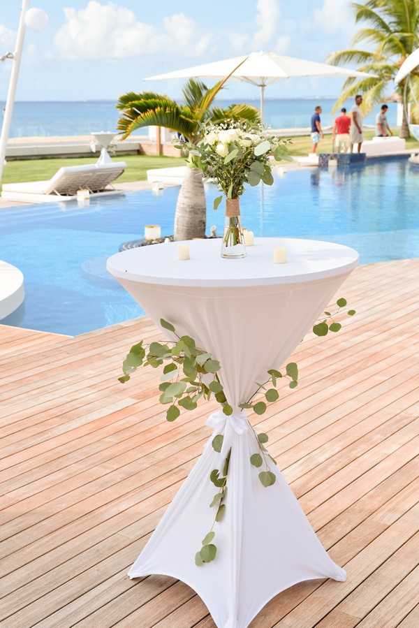 Detail shot of a cocktail hour high-top table set on a wooden deck beside a large outdoor pool with the ocean visible in the background. The round table is covered in a white spandex linen, gathered and tied at the base with trailing silver dollar eucalyptus sprigs draped around the knot. The tabletop holds a small floral arrangement in a glass vase wrapped in burlap, featuring white roses, white blooms, and green eucalyptus, accompanied by three small white votive candles. A few guests in casual attire are visible in the background near the pool, along with white market umbrellas and palm trees. The overall decor palette is white and green with a clean, garden-inspired cocktail setup. Potential venue feature image.
