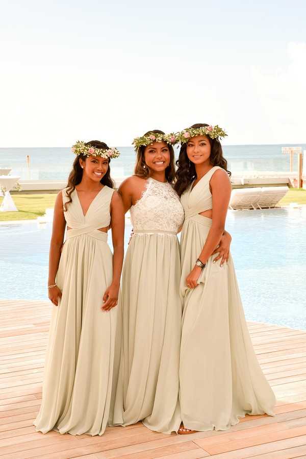 A bridal party portrait featuring three women standing on a wooden deck beside an infinity pool with the ocean visible in the background, suggesting a tropical beach venue. The bride stands in the center wearing a white halter-neck lace bodice gown with a flowing skirt and a thin belt at the waist, while the two bridesmaids wear matching sage green floor-length chiffon dresses with V-necklines and cutout waist details. All three women wear floral crowns made of greenery accented with small pink blooms. The shot is a medium portrait framed from the knees up, taken in warm natural light with a ceremony structure visible in the far background near the water's edge.