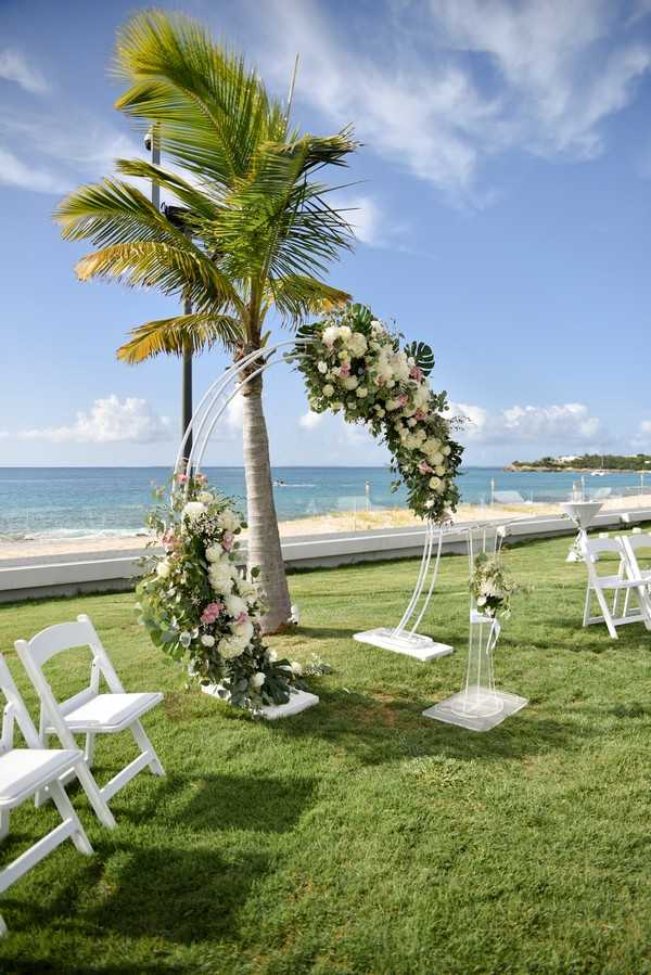 An outdoor wedding ceremony setup on a manicured lawn directly overlooking a turquoise sea, with no guests or couple present. The focal point is a circular metal arch decorated with lush floral arrangements featuring white hydrangeas, cream roses, blush pink blooms, tropical monstera leaves, and trailing eucalyptus greenery. Two smaller matching floral pedestals with acrylic stands flank the aisle entrance. White folding chairs are arranged in rows on either side of the grass aisle. The decor palette is white, blush pink, and green with a tropical-modern styling. Wide shot taken from a slight angle along the aisle.