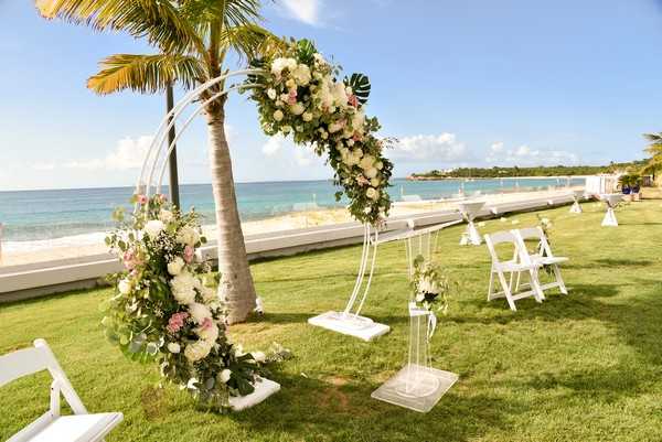 An outdoor ceremony setup on a manicured lawn overlooking the ocean, photographed in a wide shot before the ceremony begins with no guests or couple present. The focal point is a circular white metal arch decorated with a lush floral arrangement of cream and blush pink roses, white blooms, and tropical green monstera leaves with eucalyptus. A matching smaller floral arrangement adorns the base of the arch near a palm tree trunk. White folding chairs are arranged in rows on either side of a central aisle, with small floral accents on aisle-side chairs. A white acrylic or lucite podium and a small decorative white swing are positioned near the arch, adding to the clean, modern tropical aesthetic. The overall decor palette is white, cream, and blush pink with tropical greenery, consistent with a modern destination wedding style. Potential venue feature image.