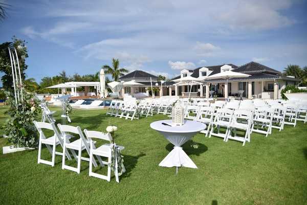 An outdoor wedding ceremony setup on a manicured lawn in front of a large white villa-style property with a dark roof, pool area, and palm trees visible in the background. White folding chairs are arranged in rows with a central aisle, and a cocktail table with a white spandex cover holds a white lantern centerpiece near the front. A floral arch on the left side of the aisle is decorated with greenery and what appear to be cream or white blooms. The overall decor palette is white and green with a clean, classic aesthetic. Wide establishing shot taken before guests have arrived. Potential venue feature image.