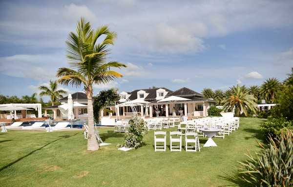 An outdoor wedding ceremony setup on a manicured lawn in front of a large white villa-style property with a dark tiled roof and wraparound covered terrace. White folding chairs are arranged in rows on either side of a central aisle, with a floral arch at the altar featuring greenery and white flowers. A cocktail-style table with a dark linen is visible near the seating area. The property includes a pool area with white umbrellas and sun loungers to the left. The wide-angle shot captures the full ceremony layout and venue grounds. Potential venue feature image.