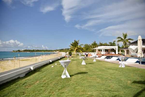 A cocktail hour or pre-reception setup is shown at an outdoor beachfront venue, with several white spandex-covered cocktail tables with small floral centerpieces arranged across a manicured lawn. The setting features a pool area with white sun loungers, a white pergola structure, and closed white market umbrellas in the background, alongside a sandy beach and turquoise water. A few guests or staff members are visible in the mid-ground near the pool area. The overall decor palette is white and minimal, consistent with a clean, modern tropical wedding aesthetic. Wide establishing shot. Potential venue feature image.