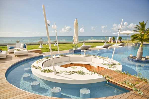 Wide-angle shot of an outdoor beachfront resort venue set up for a wedding event, with no people visible. The focal point is a circular overwater platform surrounded by a blue pool, featuring a round white cushioned lounge seating area with a wooden deck center, decorated with trailing greenery and eucalyptus garlands. Four tall white poles topped with round white globe lights frame the platform. Submerged white cylindrical stools sit in the shallow pool around the structure. The surrounding terrace includes white sun loungers, cocktail-height tables with white linens, and closed white market umbrellas. The overall decor palette is white and natural wood with green botanical accents. The ocean is visible in the background under a clear blue sky. Potential venue feature image.