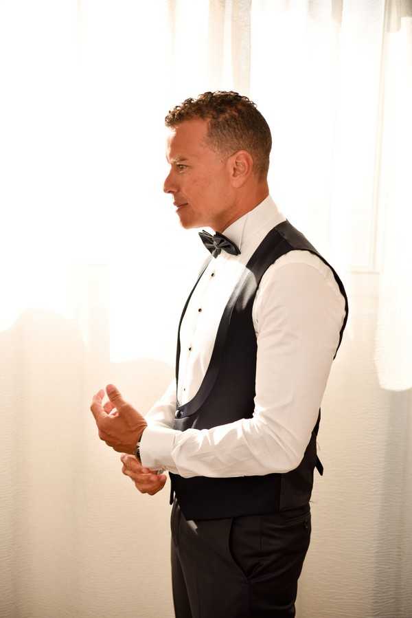 A groom getting ready, photographed in profile as he fastens his cufflinks. He is dressed in a classic black tuxedo waistcoat with satin lapels, a white formal dress shirt, and a black bow tie, paired with black trousers. The setting is an indoor room with sheer white curtains backlit by bright natural light, creating a warm, overexposed background. The shot is a mid-length portrait with a clean, minimal composition.