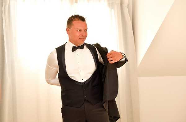A groom is captured getting ready indoors, in the process of putting on his black tuxedo jacket. He is wearing a classic black tuxedo with a matching black waistcoat, a white dress shirt with black stud buttons, and a black bow tie. The setting appears to be a bright room with white curtains and white walls. This is a medium portrait shot with the groom positioned slightly to the side, glancing over his shoulder with a relaxed expression.