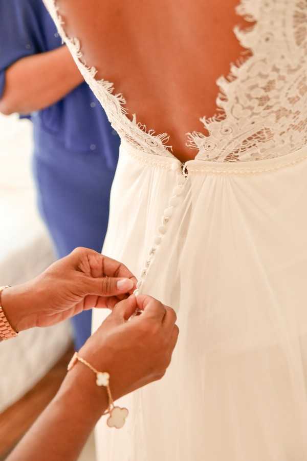 A getting-ready moment captured in close-up detail, showing a helper's hands fastening small pearl or fabric buttons up the back of a bride's white wedding gown. The dress features a deep V-back with intricate eyelash lace trim along the edges and a flowy chiffon skirt below the waist. The helper is wearing a gold chain bracelet with a clover-shaped charm, likely a Van Cleef & Arpels-style piece, and a gold watch. A second person in a blue top is partially visible in the background. The composition is a tight detail portrait shot with a softly blurred indoor background.