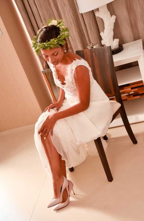 A bride is seated on a wooden chair in what appears to be a hotel room, photographed during the getting-ready phase. She is wearing a white lace-bodice wedding dress with a flowing chiffon skirt, paired with blush-white pointed-toe stiletto heels. She wears a full green leafy floral crown with a small white bloom, and pearl drop earrings, and is looking downward with her hands resting on her legs. The room features warm wood-toned furniture, beige drapery, and a white sculptural lamp in the background. The portrait-style shot is taken from a slight low angle, capturing the bride from head to toe.