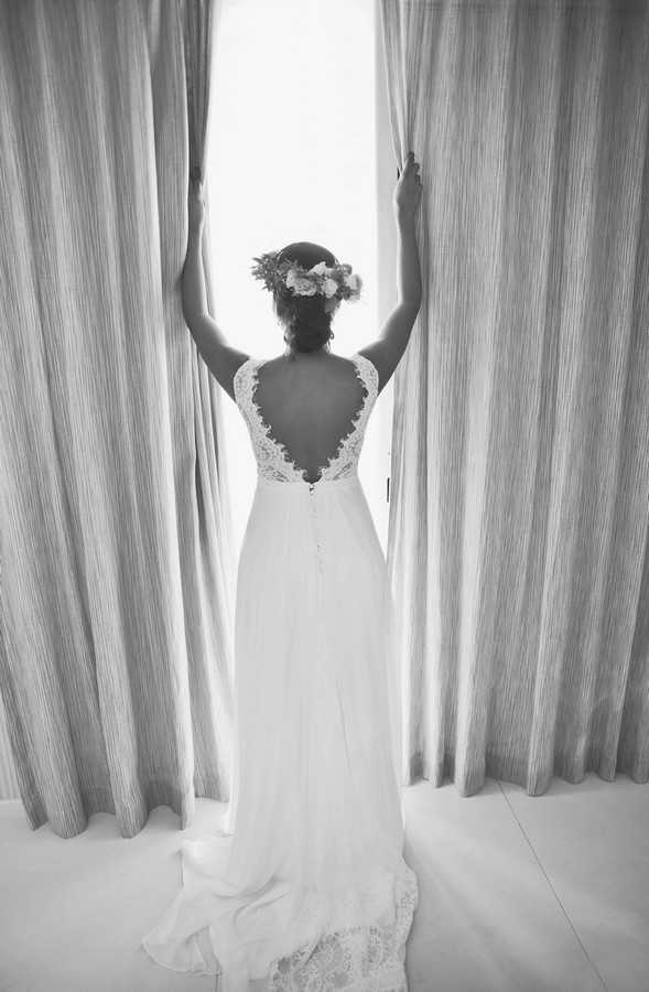 A black-and-white portrait of a bride shot from behind as she pulls open two floor-length curtains in front of a bright window, likely taken during a getting-ready moment indoors. She wears a flowing wedding gown with a deep V-back featuring scalloped lace trim and a lace hem at the base of the skirt, with thin lace shoulder straps. Her dark hair is styled in an updo adorned with a floral crown containing light-toned blooms. The image is high contrast with bright light flooding through the gap in the curtains, set against the mid-grey striped fabric of the drapes. The composition is a full-length portrait framing the bride centrally from behind.