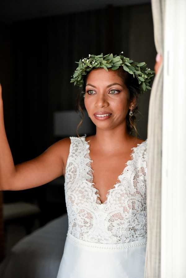 A close-up portrait of a bride during the getting-ready phase, photographed indoors against a dark background with a sheer white curtain visible at the right edge of the frame. She wears a white lace bodice gown with a deep V-neckline and a light blue satin skirt, paired with a green foliage crown made of small rounded leaves and tiny white accent buds in a boho style. Her hair is pinned up, and she wears delicate drop earrings with natural-toned makeup including defined eyes and a warm lip. She is looking slightly off-camera with a relaxed expression, and one arm is raised to hold the curtain panel.