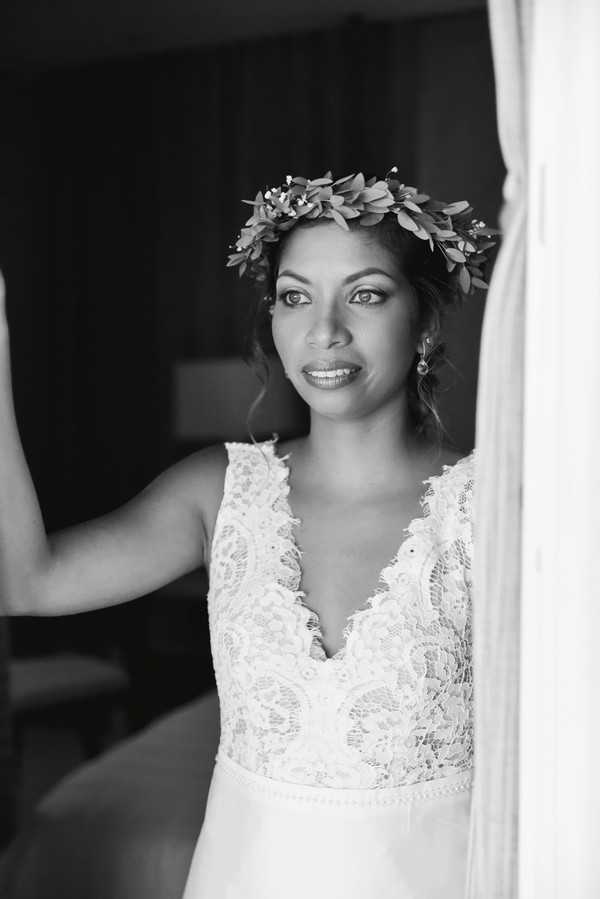 A black-and-white close-up portrait of a bride standing near a window with sheer curtains, shot with soft natural light creating gentle contrast against a dark background. She wears a deep V-neck lace bodice wedding dress with intricate floral lace detailing and a leafy greenery crown with small accent blooms positioned on her upswept hair. She wears drop earrings and looks directly into the camera with a composed expression, consistent with a getting-ready or pre-ceremony portrait.