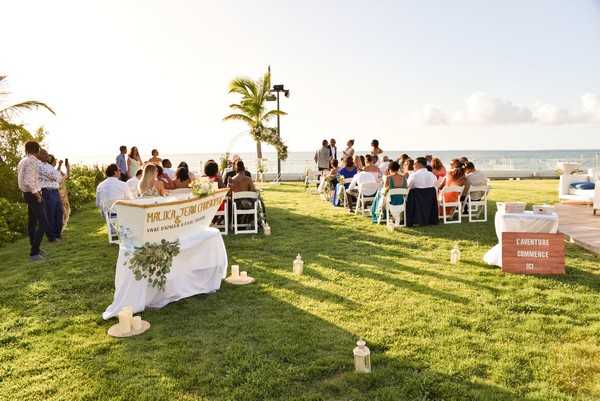 An outdoor wedding ceremony taking place on a grass lawn directly overlooking the ocean, shot as a wide angle from behind and to the side of the seating area. Approximately 30-40 guests are seated in white folding chairs arranged in rows, facing the officiant and couple at the front. In the foreground, a welcome or signing table is draped in a white cloth with a personalized banner reading 'Malika & Jean Christophe' and decorated with a greenery arrangement; white lanterns are placed on the grass nearby. A wooden sign reading 'L'Aventure Commence' is positioned to the right. The decor style is relaxed and tropical, with minimal florals and a natural color palette. The ceremony is set at golden hour, with warm low sunlight and a palm tree visible in the background against the sea horizon.