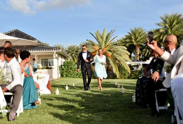 A groom in a black tuxedo with a white dress shirt and black bow tie walks down the outdoor ceremony aisle escorted by a woman in a knee-length light blue dress, likely his mother. The ceremony is set on a manicured lawn in front of a low white villa-style building with a dark tiled roof, flanked by tall palm trees. White folding chairs are arranged on either side of the grass aisle, lined with small white lanterns as aisle markers, with approximately 15–20 guests visible; a bridesmaid in a teal floor-length dress is seated on the left. The shot is a medium wide angle taken from ground level, capturing the processional moment with guests watching and one guest on the right holding up a phone to record.