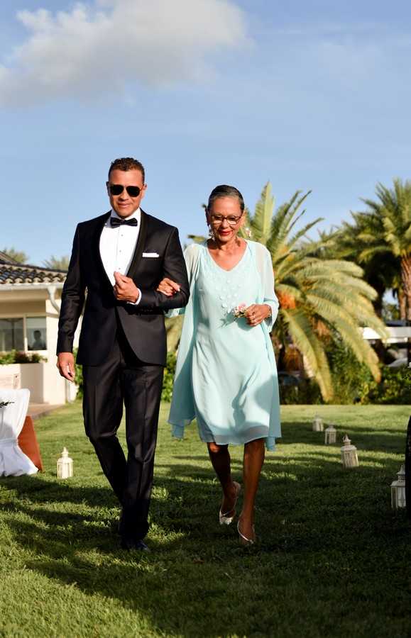 A groom in a black tuxedo with bow tie and sunglasses escorts an older woman, likely his mother, down the outdoor ceremony aisle on a grass lawn. The woman is wearing a knee-length mint green chiffon dress with a matching sheer wrap and holds a small floral wrist corsage with pale pink flowers. White lanterns line the aisle on both sides. The setting appears to be a Mediterranean-style villa or resort with palm trees and terracotta roofing visible in the background. This is a full-length portrait shot taken during the processional.