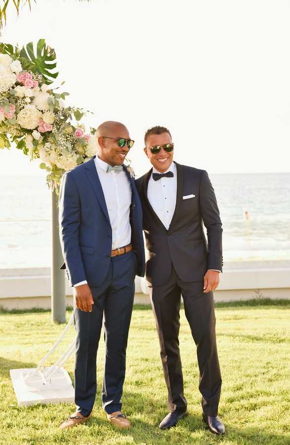 Two men pose together at an outdoor seaside wedding ceremony, both wearing sunglasses and smiling. The man on the left wears a navy blue suit with a light blue bow tie and brown leather belt, while the man on the right wears a darker navy tuxedo with a black bow tie and white pocket square. Behind them is a ceremony arch post decorated with white hydrangeas, blush pink roses, baby's breath, and tropical monstera leaves, with the ocean visible in the background. This is a portrait-style shot taken in bright natural light on a lawn area.
