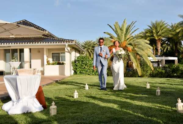 A bride and groom walk together across a manicured lawn in an outdoor garden ceremony setting, likely the processional or recessional. The groom wears a light blue suit with sunglasses, and the bride wears a fitted white gown and carries a large bouquet of white blooms and greenery. The aisle is lined on both sides with white metal lanterns placed directly on the grass, and cocktail tables draped in white linen are visible to the left. The venue is a low-rise cream-colored villa with a terracotta-tiled roof and a wooden deck visible in the background. This is a wide shot capturing the full scene with palm trees framing the background, suggesting a warm southern French or Mediterranean coastal location.