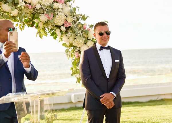 The groom stands at the altar during an outdoor ceremony, wearing a navy tuxedo with black shawl lapels, a white pocket square, and a dark bow tie, with aviator sunglasses. He is positioned in front of a floral arch decorated with white hydrangeas, blush pink roses, baby's breath, and green eucalyptus foliage. The ceremony takes place on a lawn directly overlooking the sea, visible in the background. To the left, a guest in a navy suit photographs the moment on a smartphone beside a clear acrylic lectern. Medium portrait shot.