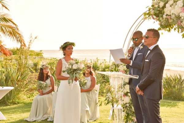 An outdoor beach wedding ceremony taking place on a garden lawn with the ocean visible in the background at golden hour. The bride wears a sleeveless white lace gown and a green leafy flower crown, holding a bouquet of white roses and eucalyptus, while the groom stands in a navy suit beside an officiant in a light blue suit who reads from papers at a clear acrylic podium. Behind the couple, two young flower girls in sage green dresses also wear small floral crowns. The ceremony arch is a circular white metal structure adorned with white and blush pink florals and lush greenery, with matching white floral arrangements on tall stands flanking the podium. The overall styling is boho-garden with a white, blush, and greenery color palette, captured in a medium wide shot from a slight side angle.