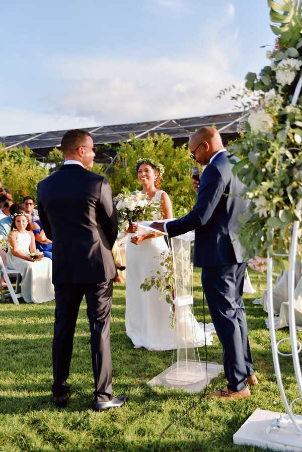 An outdoor wedding ceremony taking place on a grass lawn, with the bride and groom standing facing each other before an officiant who is speaking into a microphone at a clear acrylic lectern. The bride wears a sleeveless white gown and a green floral crown, holding a loose cascading bouquet of white flowers and eucalyptus greenery. The groom is dressed in a dark charcoal suit, while the officiant wears a navy blue suit. A white metal arch decorated with white blooms and eucalyptus foliage is partially visible on the right side of the frame. Seated guests are visible in the background, including a young flower girl in a white dress, with some guests dressed in blue and white. The setting appears to include a solar panel structure in the background. The shot is taken from a medium-wide angle, slightly behind and to the side of the groom.