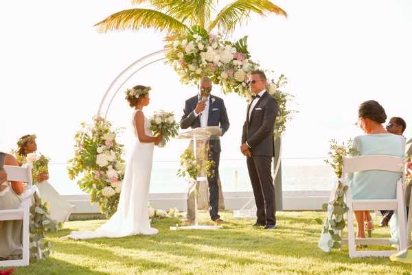 An outdoor wedding ceremony taking place on a manicured lawn with an ocean view in the background. The bride wears a sleeveless, fitted white gown and a floral crown, holding a lush bouquet of greenery and blush and white blooms, while the groom is dressed in a navy tuxedo with a black bow tie. An officiant stands between them at a clear acrylic podium beneath a circular metal arch decorated with blush pink and white florals, tropical greenery, and large leaves. Guests are seated in white folding chairs on either side of the aisle, with some women wearing light blue and beige outfits, and additional floral arrangements in blush and white with tropical foliage mark the aisle. The overall decor palette is soft blush, white, and green with a tropical-modern styling. Wide shot taken from a slight angle at ground level.
