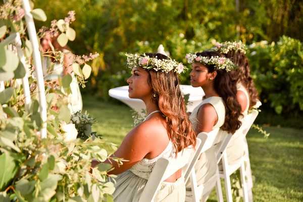 Two bridesmaids are seated in white folding chairs during an outdoor garden ceremony, viewed from a side angle. Both women wear light sage or ivory strapless dresses and floral crowns made of small pink blooms and greenery, with their long wavy hair worn down. The foreground features lush eucalyptus and foliage garland decorating the aisle or ceremony structure, with additional guests visible in white chairs behind them. The setting is a green outdoor garden with dense foliage in the background, and the overall styling is relaxed boho with a natural, garden-inspired palette.