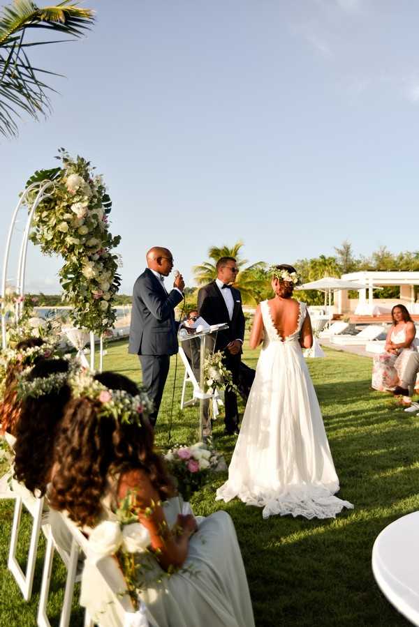An outdoor wedding ceremony taking place on a manicured lawn in a tropical resort setting, with a pool area and white cabanas visible in the background. The bride wears a white gown with a deeply cut open back and lace detailing, paired with a floral crown of white and blush blooms, while the groom wears a black tuxedo with bow tie; an officiant in a navy suit speaks at a clear acrylic podium. The ceremony arch is a curved white metal structure adorned with a lush arrangement of white and blush roses, tropical monstera leaves, and greenery. In the foreground, two young flower girls in white dresses and floral crowns are seated in white folding chairs, holding small bouquets; the shot is taken from a medium-wide angle slightly behind and to the side of the guests.