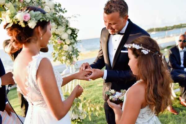 The ring exchange moment of an outdoor wedding ceremony, taking place near a waterfront with a body of water visible in the background. The groom, wearing a navy tuxedo with a black bow tie and white pocket square, places a ring on the bride's finger while smiling down at her hand; the bride wears a white lace-back gown and a large floral crown made of blush pink roses, white blooms, and greenery. A young flower girl in a white dress with a small white floral crown holds a decorative ring pillow made of succulents and greenery. A large floral arrangement of white and blush flowers with eucalyptus is partially visible in the background, and several guests are seated to the right. Medium close-up portrait shot capturing three figures in the foreground.