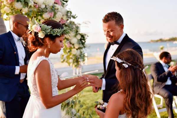 An outdoor beachside wedding ceremony showing the ring exchange moment, with the bride placing a ring on the groom's finger. The bride wears a sleeveless lace white gown and a floral crown featuring blush pink roses and greenery, while the groom is dressed in a black tuxedo with a bow tie. A young girl, likely a flower girl, stands in the foreground wearing a white floral crown and holding flowers. The ceremony arch in the background is decorated with blush pink and white blooms with lush greenery. Several guests are visible seated in white chairs to the right, and a male officiant in a navy suit stands behind the couple. The image is a medium close-up portrait shot taken in warm late-afternoon light.
