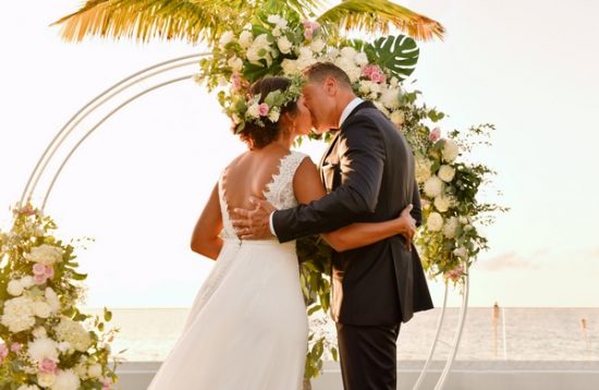 The bride and groom share their first kiss during an outdoor ceremony set directly in front of the ocean at what appears to be a beachfront venue. The couple stands beneath a circular gold hoop arch decorated with white hydrangeas, pink roses, monstera leaves, and trailing greenery. The bride wears a white A-line gown with a lace open back and a floral crown featuring pink and white blooms in her hair, while the groom wears a dark navy suit. The composition is a medium portrait shot taken from slightly behind and to the side of the couple, with the ceremony arch and ocean horizon visible in the background.