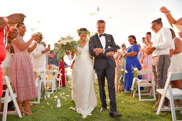 A bride and groom walk back down the aisle during a ceremony recessional at an outdoor wedding, with approximately 20 guests lining both sides throwing white rose petals. The ceremony takes place on a lawn with white folding chairs arranged in rows, and the aisle chairs are decorated with small white floral arrangements. The bride wears a slim ivory gown with a green foliage crown and carries a large bouquet of white flowers with lush tropical greenery; the groom wears a dark navy suit with a black bow tie and white pocket square. Guests are dressed in a range of colorful outfits including dusty pink lace, cobalt blue, and white. The image is a wide-angle shot taken at golden hour, with warm late-day sunlight illuminating the scene from behind the couple.