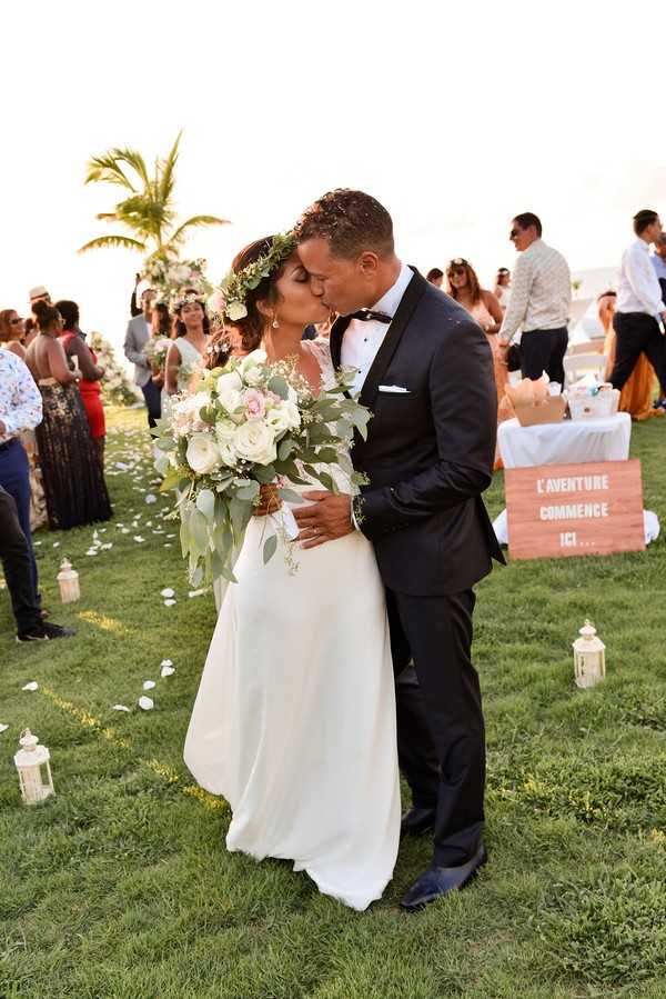 The bride and groom share their first kiss at the end of the ceremony aisle during an outdoor wedding on a tropical lawn setting, with a palm tree and ocean horizon visible in the background. The bride wears a simple ivory crepe gown and a lush floral crown of white and green foliage, carrying an oversized cascading bouquet of white roses, blush pink roses, and eucalyptus; the groom wears a navy tuxedo with a black bow tie. White lanterns line the grass aisle, scattered with white flower petals, and a wooden sign reading 'L'AVENTURE COMMENCE ICI...' is visible to the right alongside a white-clothed table. Approximately 20 guests are visible in the background watching the ceremony, and the shot is a mid-length portrait taken at ground level with the couple as the sharp focal point against a softly blurred crowd.