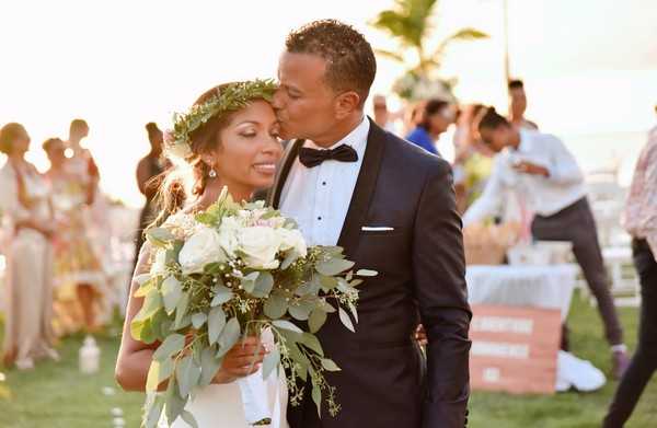 The bride and groom share a kiss in a close-up portrait shot taken outdoors, likely following a garden or beachside ceremony. The bride wears a white gown and a green eucalyptus leaf crown, and holds a loose bouquet of ivory roses, white blooms, and trailing eucalyptus foliage. The groom is dressed in a navy tuxedo with a black bow tie and white pocket square. In the soft-focus background, a group of guests in summery attire mingles on a lawn, with palm trees and warm golden-hour light visible.