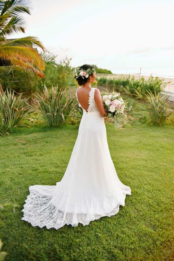 A bridal portrait taken outdoors in a garden setting, showing the bride from behind as she faces away from the camera. She wears a white A-line gown with a deep V-back featuring lace detailing and a flowing train edged with lace. Her dark hair is styled in an updo adorned with a floral crown of white and blush blooms with greenery. She holds a loose bouquet of blush and white roses with eucalyptus and mixed greenery. The composition is a full-length portrait emphasizing the back of the dress and train spread across a lawn, with tropical and Mediterranean-style plantings visible in the background. The overall styling is boho-romantic.