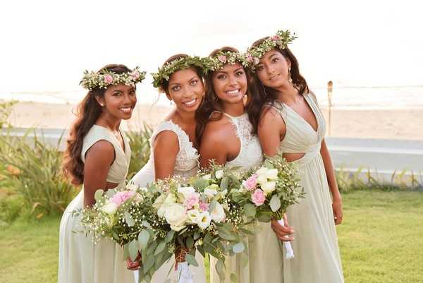 A bridal party portrait featuring a bride and three bridesmaids posing outdoors near a beach, with sand and ocean visible in the background. The bride wears a white two-piece lace crop top and skirt ensemble, while the three bridesmaids wear sage green floor-length dresses in a wrap/draped style. All four women wear floral crowns made of greenery, small pink blooms, and baby's breath. They each hold bouquets composed of ivory calla lilies, blush and pink roses, white ranunculus, baby's breath, and trailing eucalyptus greenery. The overall color palette is soft sage, blush, and white with a relaxed, boho-inspired aesthetic. The shot is a close-up group portrait taken at golden hour with warm, diffused natural light.
