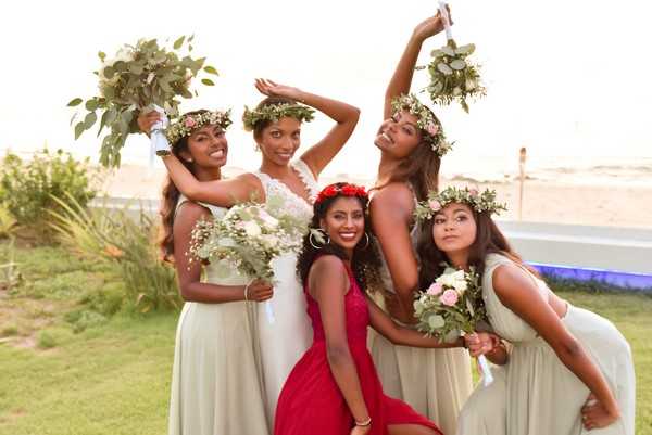 A bridal party portrait taken outdoors near a beach, featuring the bride and four attendants posing playfully together. The bride wears a white lace gown and a green eucalyptus flower crown, while three bridesmaids wear sage/light grey floor-length dresses and matching eucalyptus and small floral crowns; one attendant wears a red dress and a red floral crown. The bouquets are composed of white and blush pink roses, eucalyptus, and baby's breath. The group is mid-pose with dynamic, energetic positioning, and a sandy beach and blue pool structure are visible in the background. The shot is a close-up group portrait with a warm, bright ambient light.