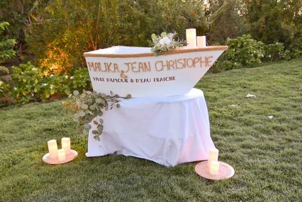 A decorative wedding display set outdoors on a lawn, featuring a white wooden boat prop personalized with the text 'MALIKA & JEAN CHRISTOPHE' and the phrase 'VIVRE D'AMOUR & D'EAU FRAÎCHE' painted in gold lettering on its side. The boat is placed on a white-linen-draped round table and is adorned with white candles and a small arrangement of white blooms and trailing eucalyptus. Four flameless pillar candles in pink/rose-tinted holders are placed on the grass around the base of the table. The shot is a medium wide angle capturing the full decorative vignette against a backdrop of garden greenery at dusk.