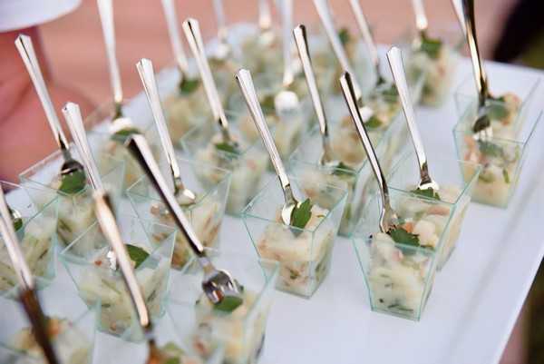 Close-up detail shot of a cocktail hour canapé display featuring rows of small clear square plastic cups filled with a pale green seafood or vegetable appetizer garnished with fresh herb leaves, each cup fitted with a small silver cocktail fork or spoon. The cups are arranged in uniform rows on a white surface, suggesting an outdoor or covered reception setting. The styling is clean and modern, with the clear vessels and polished silver utensils reflecting a classic catering presentation. Approximately 20 or more individual portions are visible in the frame.