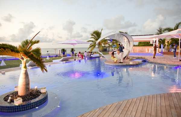 A cocktail hour or reception gathering takes place on an outdoor pool terrace at an oceanfront venue, photographed at dusk with a wide shot composition. The pool features blue underwater LED lighting, a small island with a palm tree at its center, and a large spherical pod-style seating structure on an island within the pool where two guests are seated. Approximately 10–15 guests are visible mingling across the terrace, with white canopy structures and pink parasols providing shade in the background. The decor palette is modern and minimalist, with white furnishings and cool blue lighting contrasting against the warm dusk sky and ocean backdrop. Potential venue feature image.