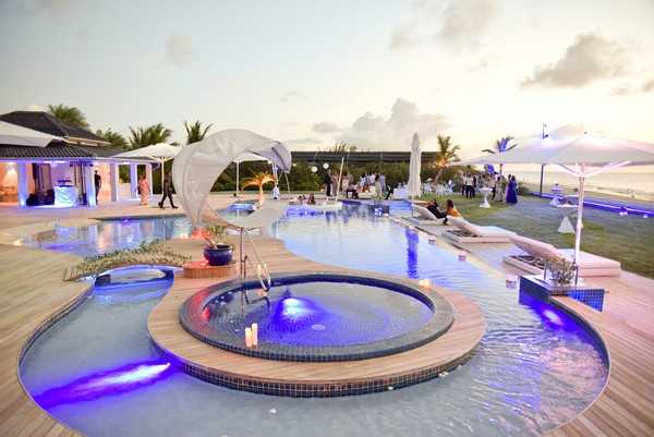 A wide-angle shot of an outdoor cocktail hour or reception taking place on a large pool terrace at dusk, with the ocean visible in the background. The venue features a freeform swimming pool with an illuminated hot tub/jacuzzi in the foreground lit in deep purple and blue, surrounded by wooden decking. Guests dressed in formal and semi-formal attire — including some in white and others in dark clothing — are mingling across the terrace, which is furnished with white lounge chairs, white market umbrellas, and a draped canopy structure. The main building to the left is warmly lit with warm amber lighting under a covered pergola or pavilion. The overall decor palette is white and blue with modern, clean styling. Potential venue feature image.