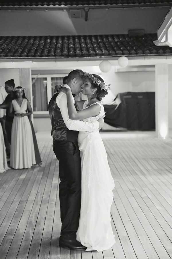 This black-and-white image captures the couple's first dance on a wooden deck beneath a covered outdoor terrace with a tiled roof. The bride wears a flowing, lightweight white gown with lace detailing at the shoulders and a floral crown in her upswept hair, while the groom is dressed in dark trousers, a waistcoat, and a dress shirt. The couple is embraced closely, the bride smiling up at the groom, with soft contrast between their light and dark tones. In the background, a bridesmaid in a long flowing dress and a floral crown is visible, along with at least one other guest, and round paper lanterns hang overhead providing ambient lighting. The composition is a medium full-body portrait shot focused on the couple, with the background slightly out of focus.