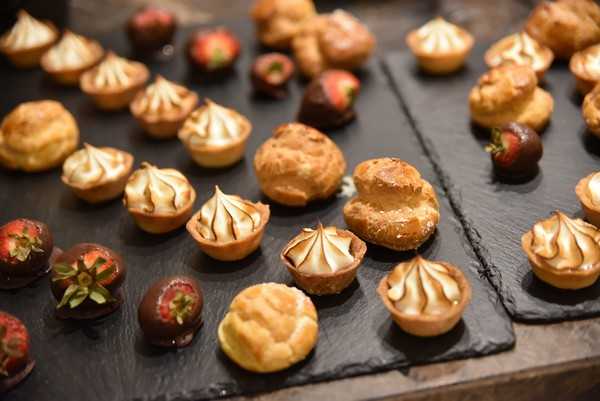 Close-up detail shot of a wedding dessert display arranged on dark slate boards. The spread includes mini lemon meringue tartlets with toasted golden-brown meringue peaks, cream puffs and choux pastry buns, and chocolate-dipped strawberries with green stems. The desserts are arranged in loose rows across multiple slate serving boards, with warm ambient lighting highlighting the caramelized meringue tones and dark chocolate coating. The overall palette is warm amber, dark brown, and deep chocolate against the charcoal slate surface.