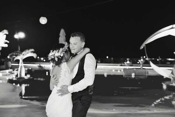 A black-and-white nighttime portrait of a couple sharing a close embrace, likely during a first dance or a quiet moment at an outdoor evening reception. The bride wears a fitted lace gown and has a floral hair piece, while the groom is dressed in a white dress shirt, dark vest, and bow tie. The background shows a poolside or waterfront terrace setting with scattered point-light sources — globe lights, lampposts, and bokeh reflections on water — creating strong contrast against the dark sky. The image is a mid-shot with soft focus on the background, drawing attention to the couple in the foreground.