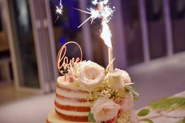 Close-up detail shot of a two-tier naked wedding cake displayed during an indoor reception. The cake features minimal frosting with visible sponge layers, decorated with blush and ivory roses, white gypsophila, and green foliage. A lit sparkler is mounted on top alongside a rose gold laser-cut 'love' cake topper. The background is softly blurred, revealing purple ambient uplighting and large glass doors or windows typical of a modern venue interior.
