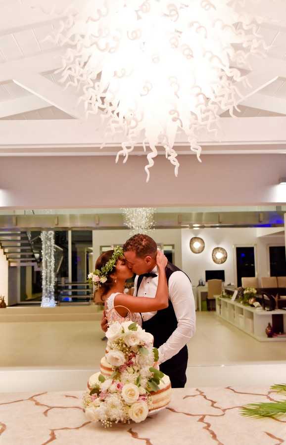 A bride and groom share a kiss during the cake cutting moment inside a modern, contemporary reception venue. The bride wears a lace wedding dress and a floral crown with white and blush blooms and greenery, while the groom is dressed in a white dress shirt, black bow tie, and black vest. In the foreground, a naked-style tiered wedding cake is heavily adorned with white roses, blush flowers, and eucalyptus greenery. Above the couple hangs a large white sculptural glass chandelier with swirling tendrils, dramatically lit from within. The interior space features mirrored walls, recessed lighting, and a staircase with fairy light decoration visible in the background. The shot is a medium portrait taken at eye level, with the cake in the lower foreground framing the couple.