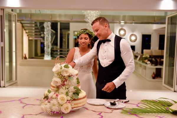 The bride and groom are cutting their wedding cake at an indoor reception in a modern, open-plan venue with white walls, a glass staircase, and contemporary lighting including a crystal chandelier in the background. The bride wears a sleeveless white lace gown and a floral crown made of greenery and small white flowers, while the groom is dressed in a white dress shirt, black vest, and bow tie. The naked-style wedding cake is decorated with white roses, pink spray roses, and trailing eucalyptus foliage. The shot is a medium portrait framing both subjects from the waist up, with the cake prominently in the foreground on a white table.