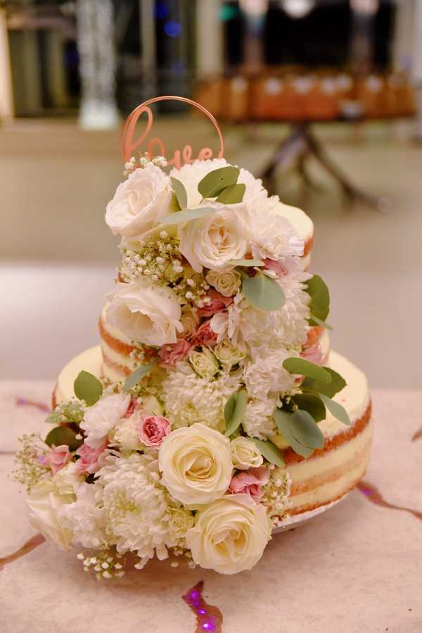 Close-up detail shot of a three-tiered naked wedding cake displayed on a table at an indoor reception venue. The cake has minimal frosting on the exterior, revealing the sponge layers, and is heavily decorated with cascading fresh flowers including cream roses, blush pink spray roses, white chrysanthemums, baby's breath, and eucalyptus leaves. A rose gold wire cake topper reading 'love' is placed at the top. The floral palette is soft, combining cream, blush pink, and white blooms with green foliage throughout all three tiers. The background is softly blurred but suggests an indoor reception space with warm lighting.