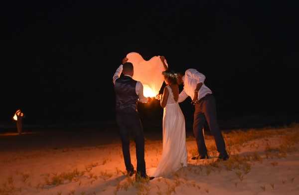 A nighttime outdoor scene on a sandy beach where a couple, assisted by two others, is releasing a large heart-shaped sky lantern. The bride wears a white flowing gown with a floral crown, and the groom is dressed in a dark suit with a waistcoat. The glowing orange flame inside the paper lantern illuminates the group from below, casting warm light against the otherwise dark surroundings. A fifth figure holding a torch is visible in the background to the left, and the overall composition is a wide shot taken from behind the group.
