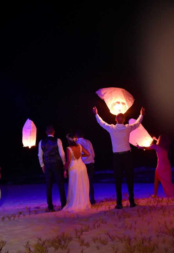 A group of wedding guests and the couple are releasing white paper sky lanterns on a sandy beach at night, with at least three glowing lanterns visible — one being held aloft with arms raised, one already airborne to the left, and one being lit to the right. The bride wears a white floor-length gown with a low open back and lace detailing, with a floral hair accessory, while the groom stands beside her in a dark waistcoat and trousers with a white shirt. The scene is lit by a mix of purple-blue ambient lighting from below and the warm orange glow of the lantern flames. Wide shot taken from behind the group, capturing the full scene across the beach setting.